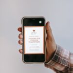 Crop anonymous person holding smartphone in hand and demonstrating screen with app with invitation to be volunteer on gray background
