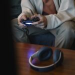 Crop unrecognizable man with joystick playing video game while sitting against table with modern headset in house room