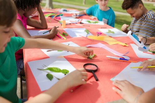 Kids engaging in fun arts and crafts, using leaves and paper outside at a colorful table.